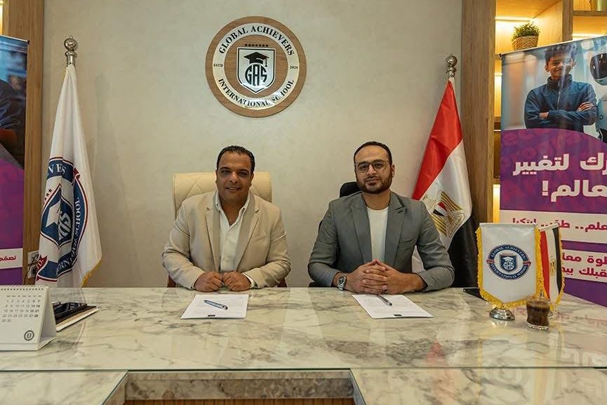 Two men seated at a marble desk with papers, below the Global Achievers International School emblem