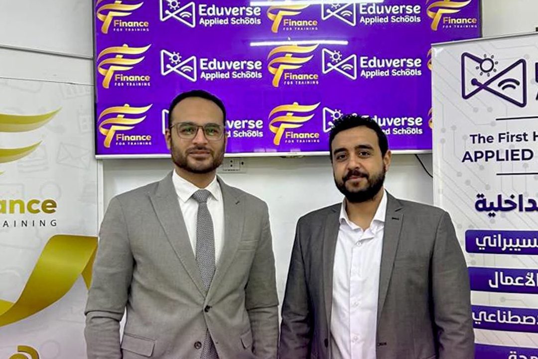 Two men in suits standing in front of a Finance for Training Academy and Eduverse Applied Schools backdrop, beside a roll-up banner