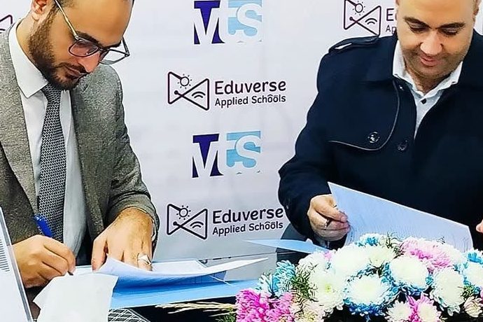 Two men signing an agreement at a table with flowers, in front of an MTS and Eduverse Applied Schools backdrop