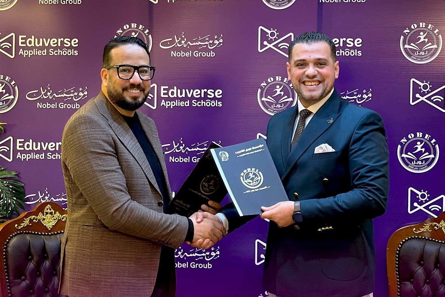 Two men shaking hands and holding folders, in front of a Nobel Group and Eduverse Applied Schools backdrop