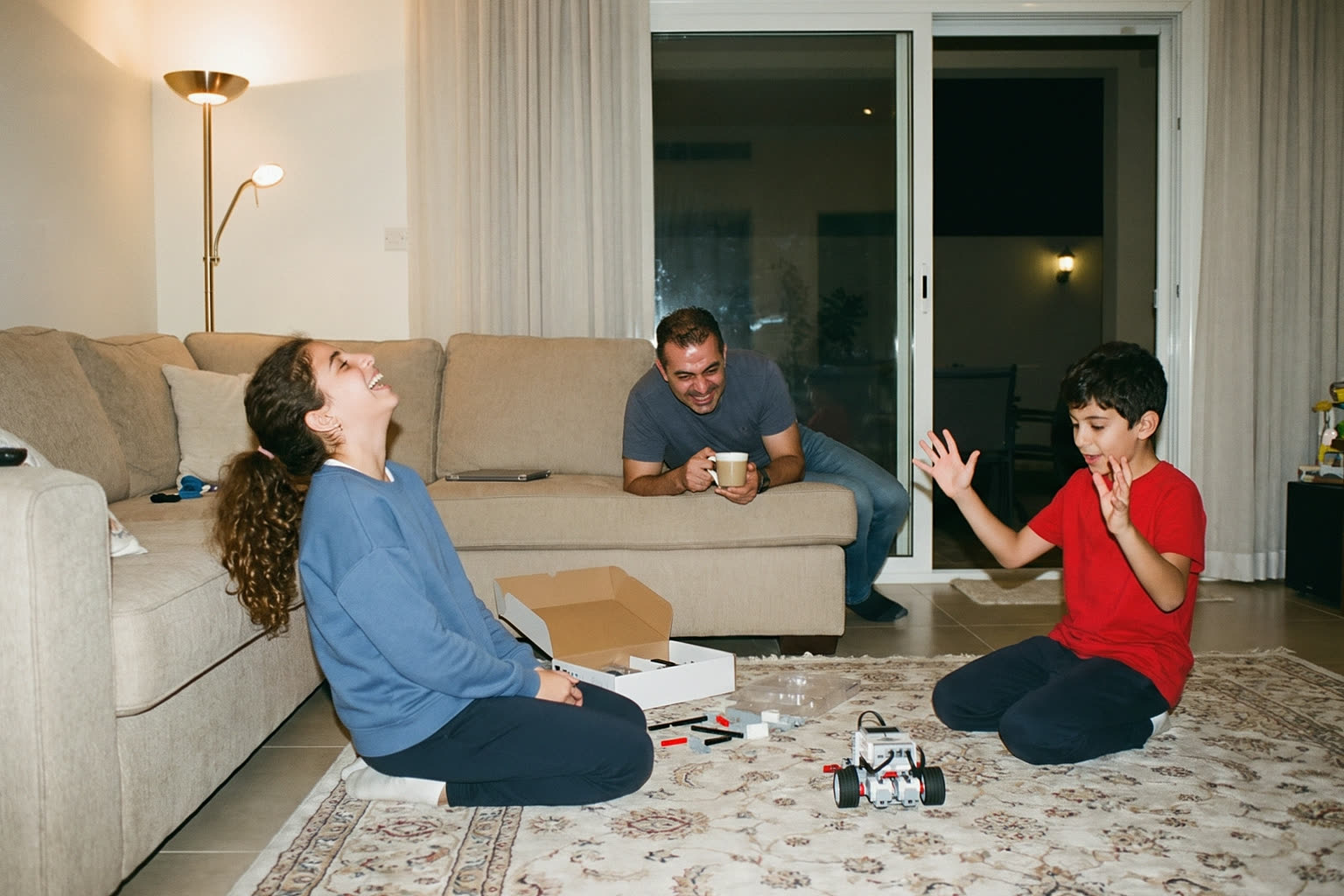 A brother and sister laughing as the robot they built rolls across the living-room rug, their father watching from the sofa